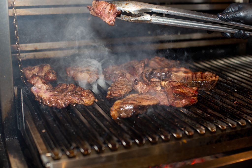 Close-up of steaks grilling on a barbecue, showcasing smoke and tongs in action.